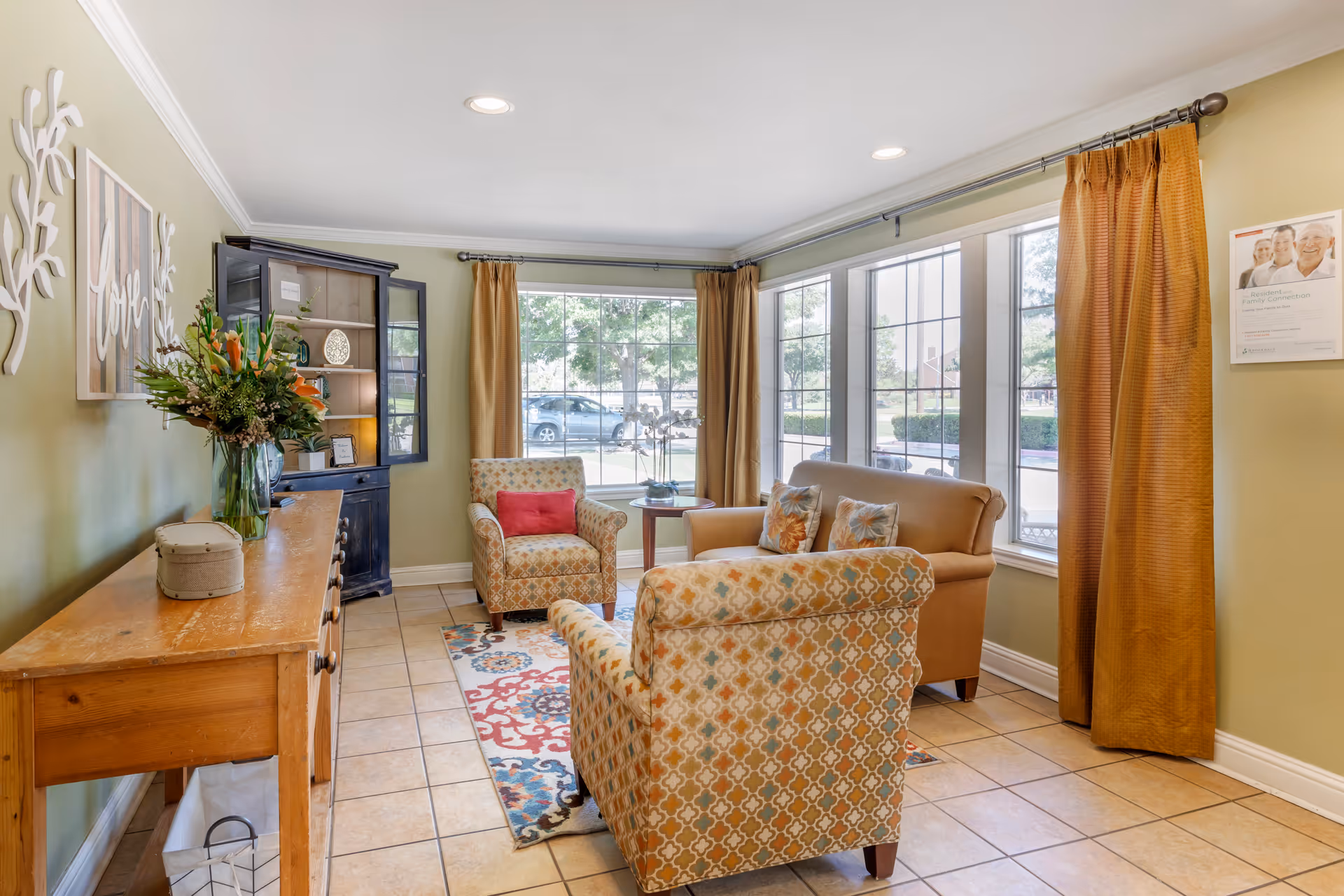 A cozy living room area with two patterned armchairs, a beige loveseat with floral pillows, a wooden side table, and a wooden console table with a vase of flowers. Large windows with brown curtains allow natural light to fill the room. The walls are painted light green and decorated with a 'love' wall art and a framed poster.