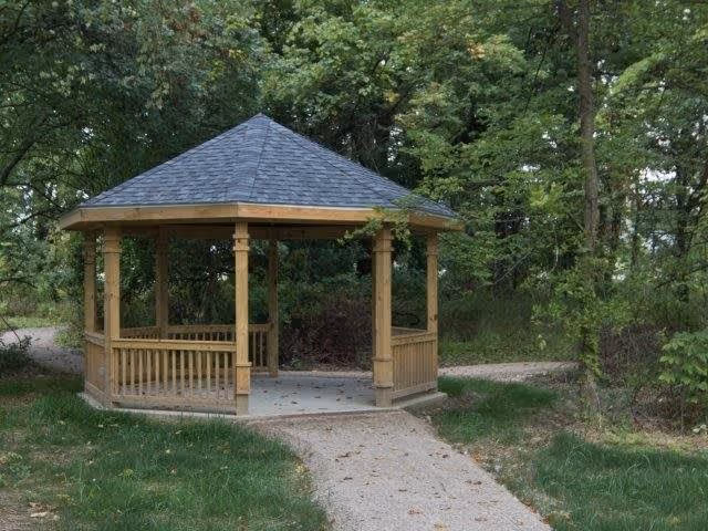 A wooden gazebo with a shingled roof situated in a green outdoor area surrounded by trees and grass, with a paved pathway leading to it.