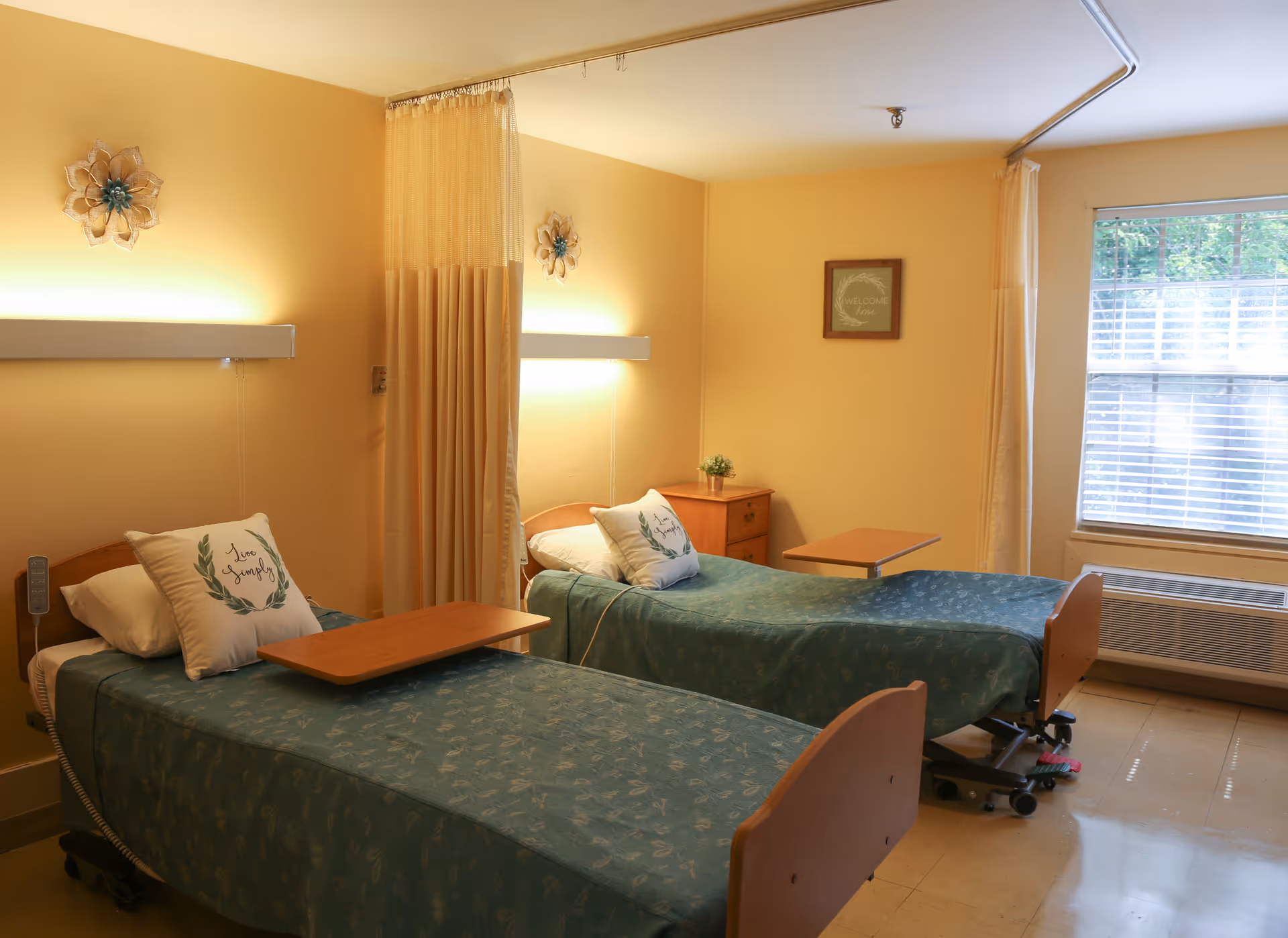 A softly lit shared patient bedroom with two hospital-style beds, green bedspreads, a privacy curtain, bedside tables and a window.