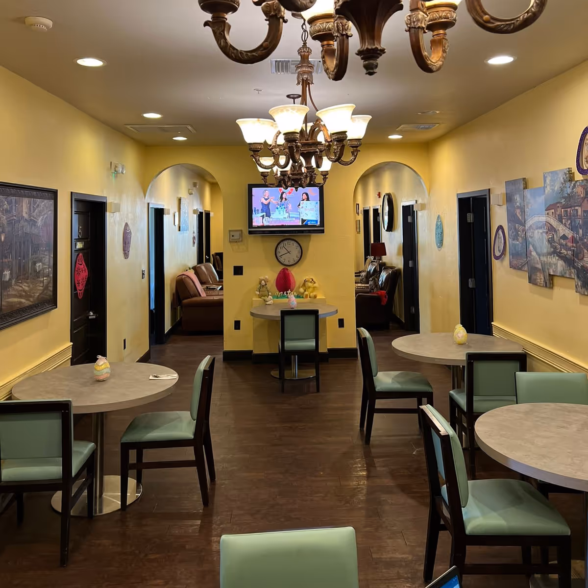Interior view of a communal dining and sitting area in an assisted living facility. The room has yellow walls, dark wood flooring, and several round tables with green cushioned chairs. There are two archways leading to seating areas with brown leather chairs. A wall-mounted TV and a clock are visible on the far wall, along with decorative items on the tables and walls. A chandelier hangs from the ceiling.