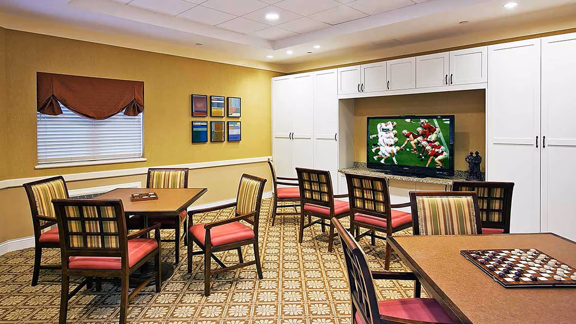 Community sitting room with multiple tables and chairs, a wall-mounted TV showing a football game, and a checkerboard on a table.