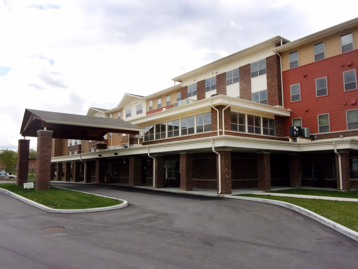 Exterior view of a multi-story senior living facility named Oasis At 30th, featuring a covered entrance with brick pillars and a driveway leading up to it. The building has a combination of brick and siding with multiple windows and a partly cloudy sky in the background.