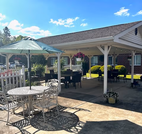 Outdoor patio area with a white metal table and chairs under a green umbrella, additional tables and chairs under a covered pavilion, hanging flower baskets, and a brick building in the background under a blue sky with some clouds.