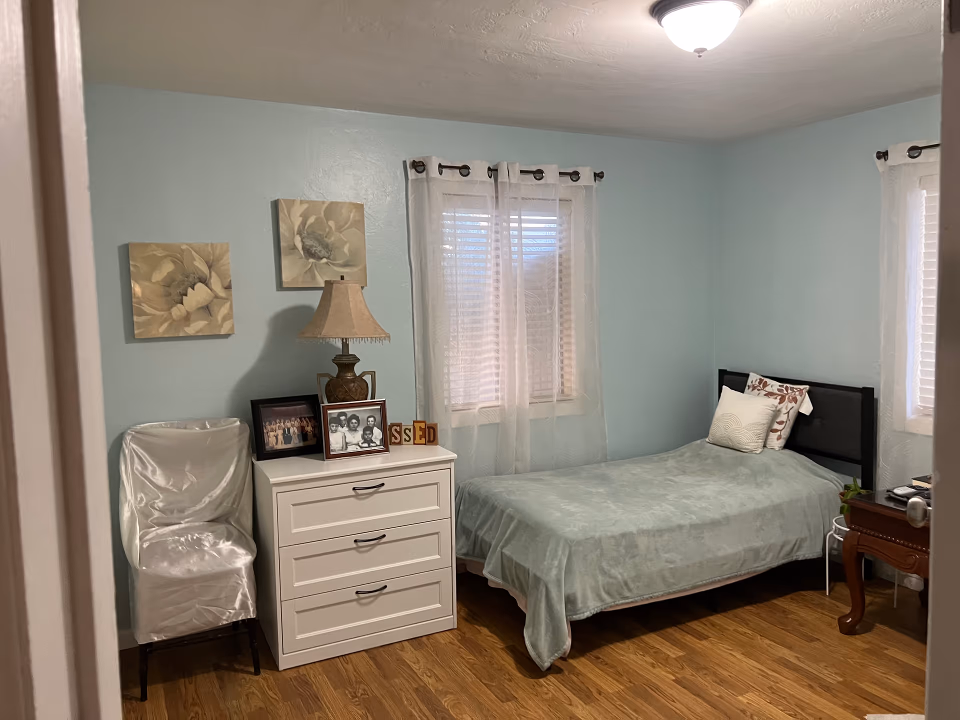 Tidy bedroom with light blue walls, single bed, dresser, chair, and sheer-curtained windows.