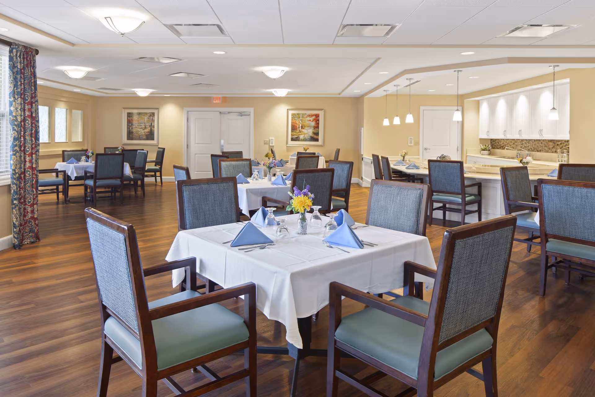 A dining room in a senior living facility with several tables covered in white tablecloths, each set with blue folded napkins, glassware, and small flower arrangements. The room has wooden floors, cushioned chairs with wooden frames, and soft yellow walls. There are framed pictures on the walls and a kitchen area with pendant lights and white cabinets in the background.