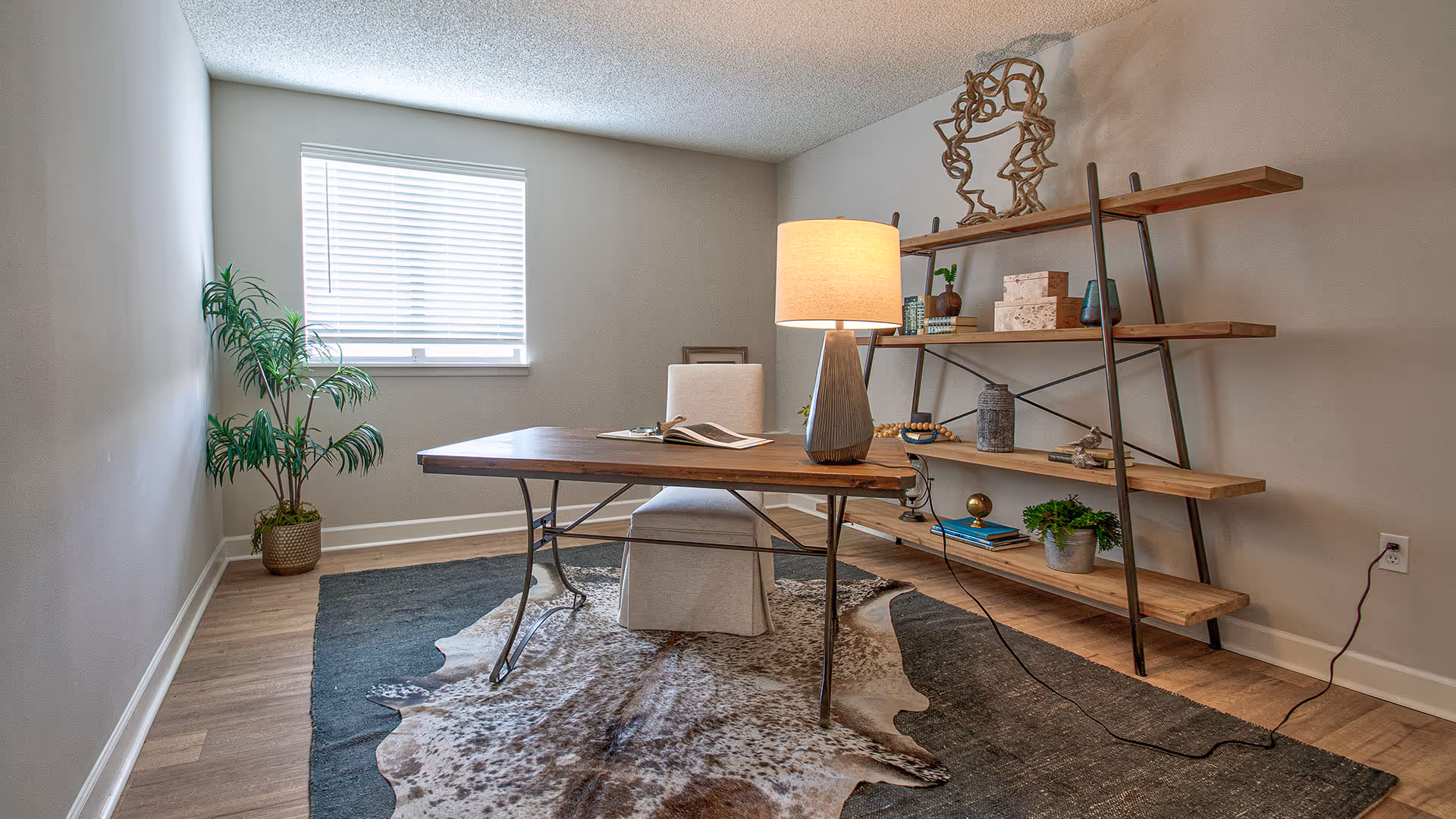 A well-lit room with a wooden desk and a beige upholstered chair. On the desk is a lit table lamp and an open book. Behind the desk is a wooden shelving unit with decorative items including plants, boxes, and sculptures. A large window with blinds is on the left wall, and a potted plant is placed near the window. The floor has a dark area rug with a cowhide rug layered on top.