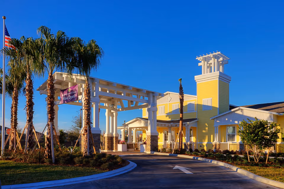 Exterior view of a senior living facility named Serenades at West Orange, featuring a yellow building with white trim, a covered entrance supported by white columns, palm trees, and a clear blue sky.