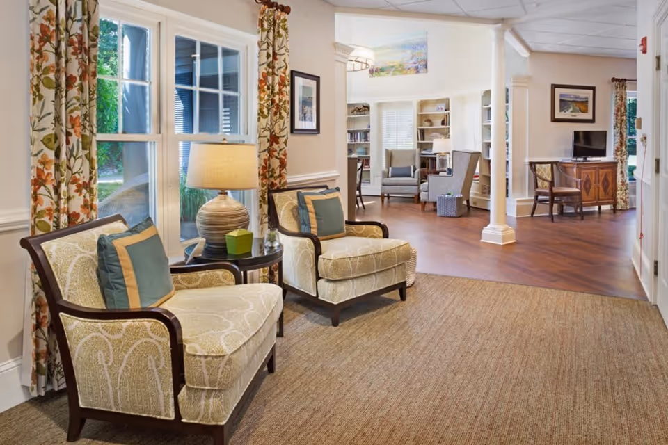 A cozy and well-lit living room area in a senior living facility featuring two upholstered armchairs with decorative pillows, a side table with a lamp, floral curtains on the windows, and an open space leading to another seating area with armchairs, bookshelves, and a TV on a wooden cabinet.