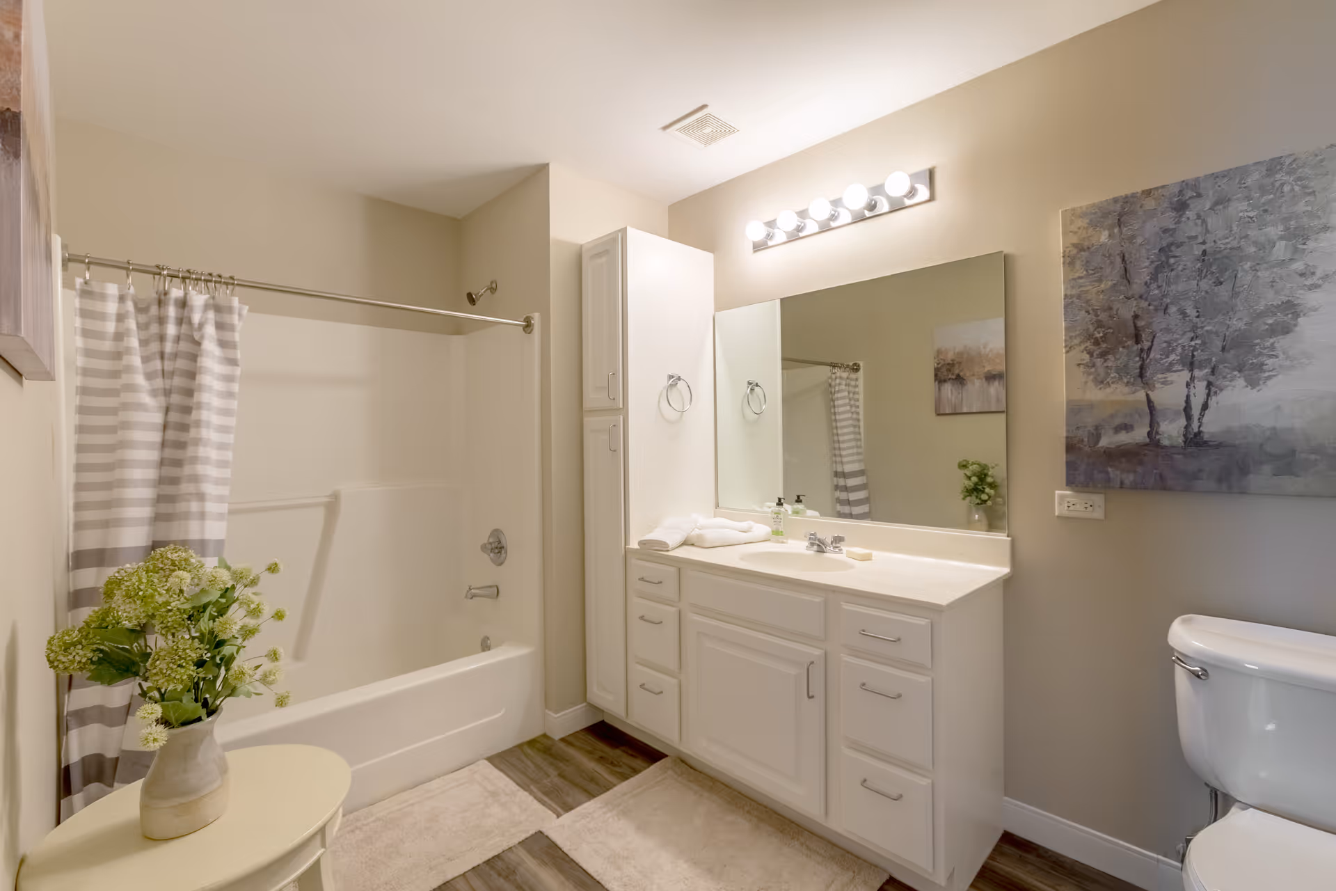 Bright, modern bathroom featuring a tub/shower with a striped curtain, a white vanity and mirror, a toilet, and wall artwork.