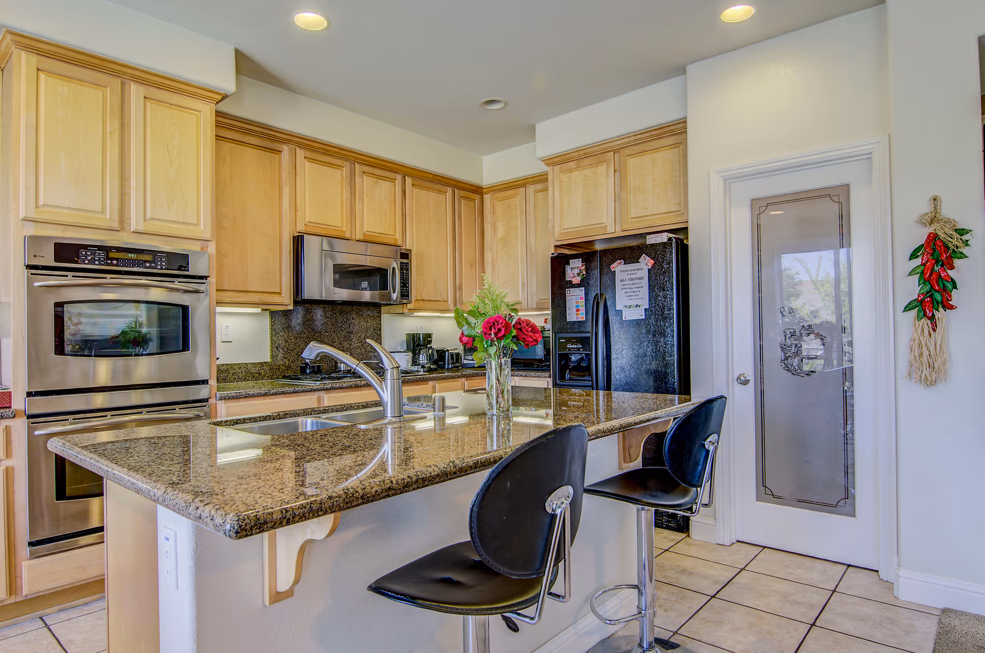 A modern kitchen with light wood cabinets, granite countertops, and stainless steel appliances including a double oven and microwave. There is a black refrigerator with notes attached, a kitchen island with a sink and two black bar stools, and a vase with red flowers on the island. A door with frosted glass and decorative etching is visible, along with a hanging decoration of red chili peppers on the wall.