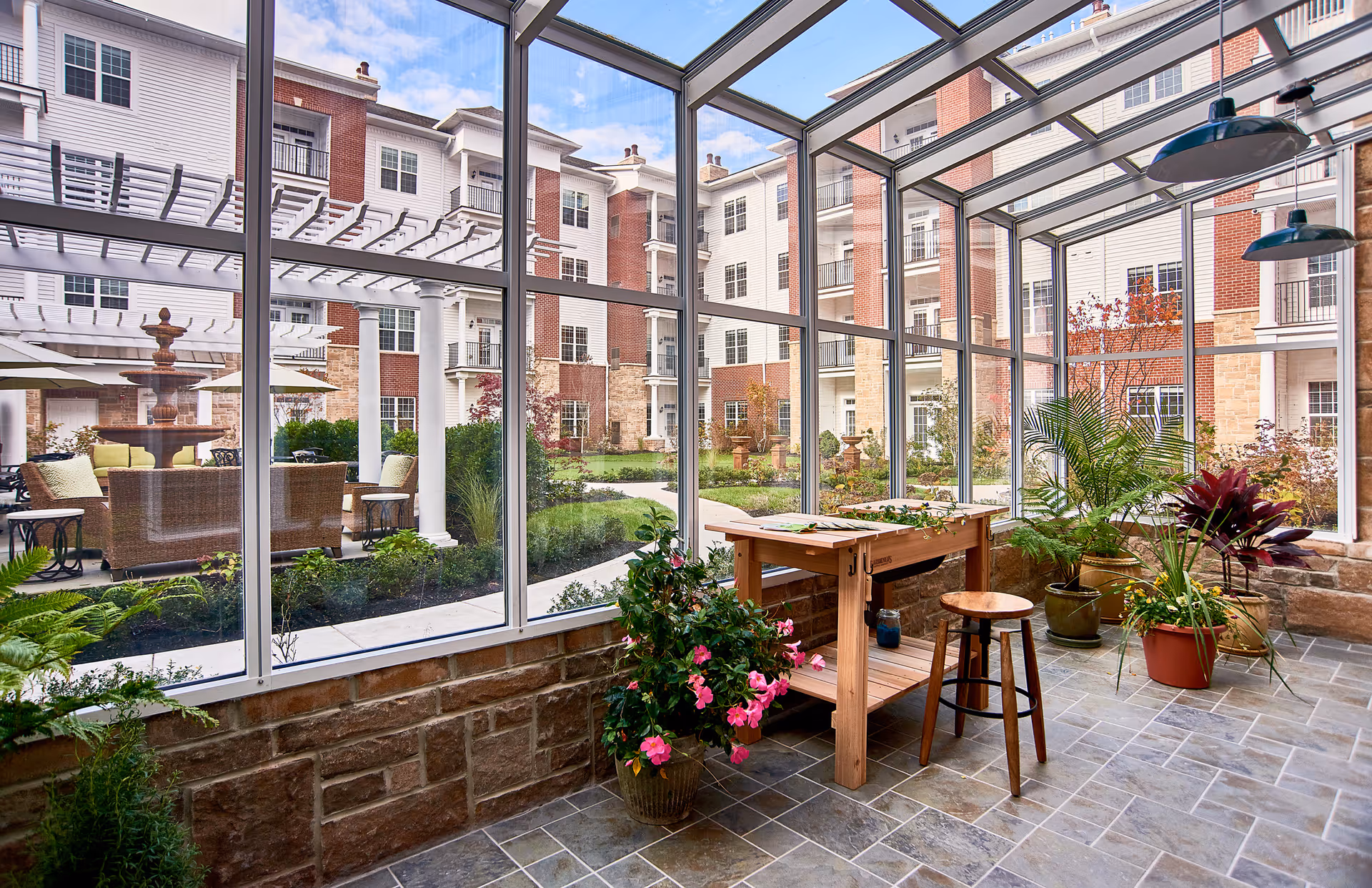 A bright indoor garden room with large glass windows and ceiling overlooking an outdoor courtyard with a fountain and seating area. Inside, there is a wooden potting bench with a stool and several potted plants on a tiled floor.