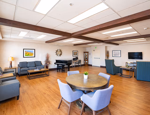 Spacious retirement community common room with seating areas, a round table and chairs, a piano, TV, and wood flooring.