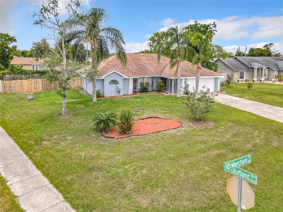 Single-story suburban house with a large front lawn, palm trees, and a driveway at a street corner.