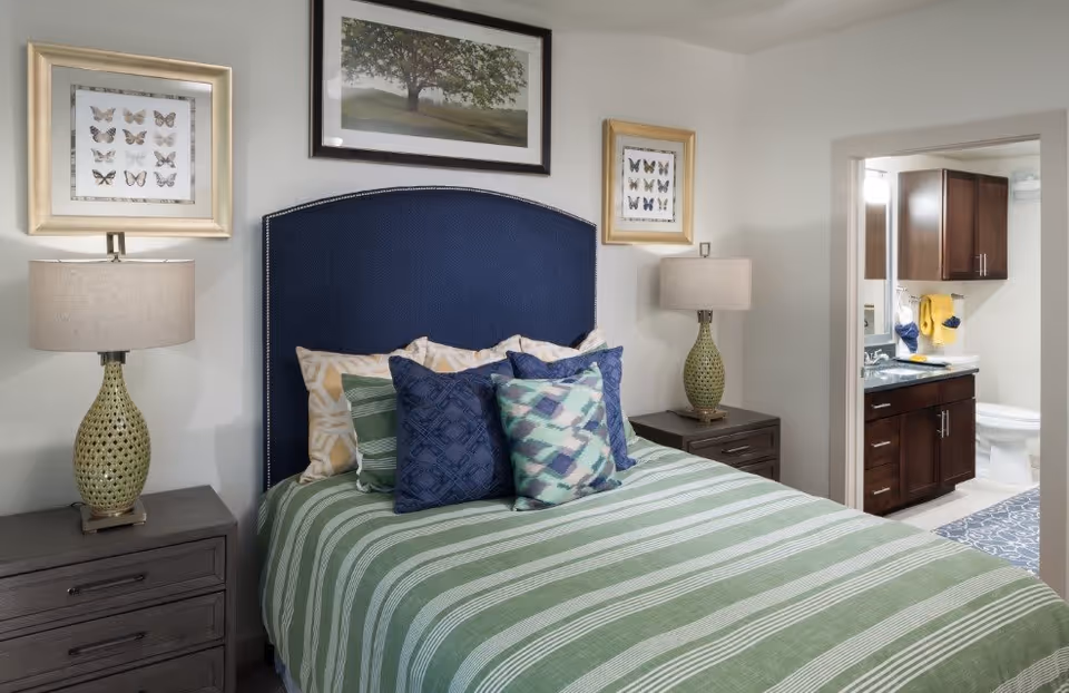 A bedroom with a bed featuring a blue upholstered headboard and green striped bedding with multiple decorative pillows. There are two bedside tables, each with a lamp and framed butterfly artwork above. A doorway leads to a bathroom with dark wood cabinets, a sink, and a toilet visible.