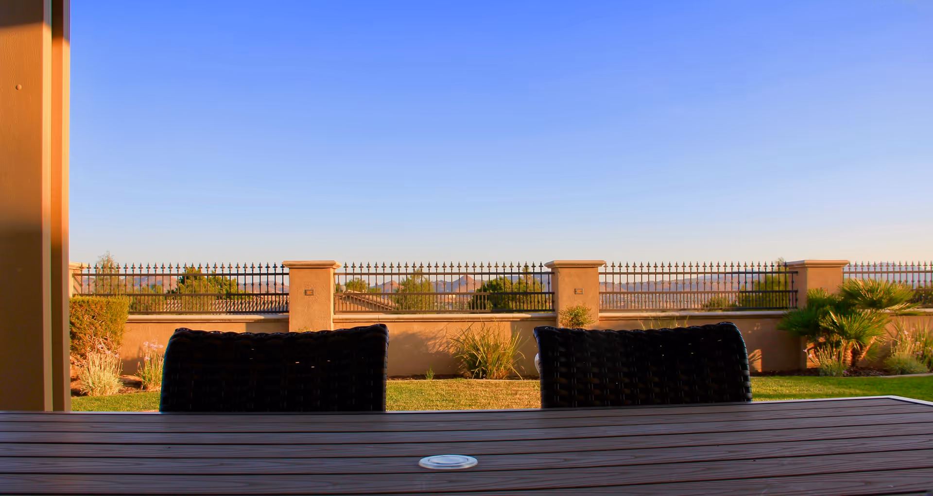 View from a covered patio area showing a wooden table with two black woven chairs. Beyond the patio is a grassy yard with some plants and shrubs, a beige wall with decorative iron fencing, and a clear blue sky above.