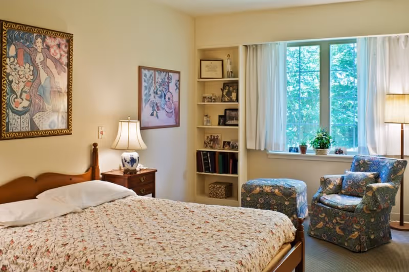 A cozy bedroom with a wooden bed covered in a floral quilt, a bedside table with a lamp, framed artwork on the walls, a built-in shelf with photos and books, a window with white curtains letting in natural light, and a floral upholstered armchair with a matching ottoman next to a floor lamp.