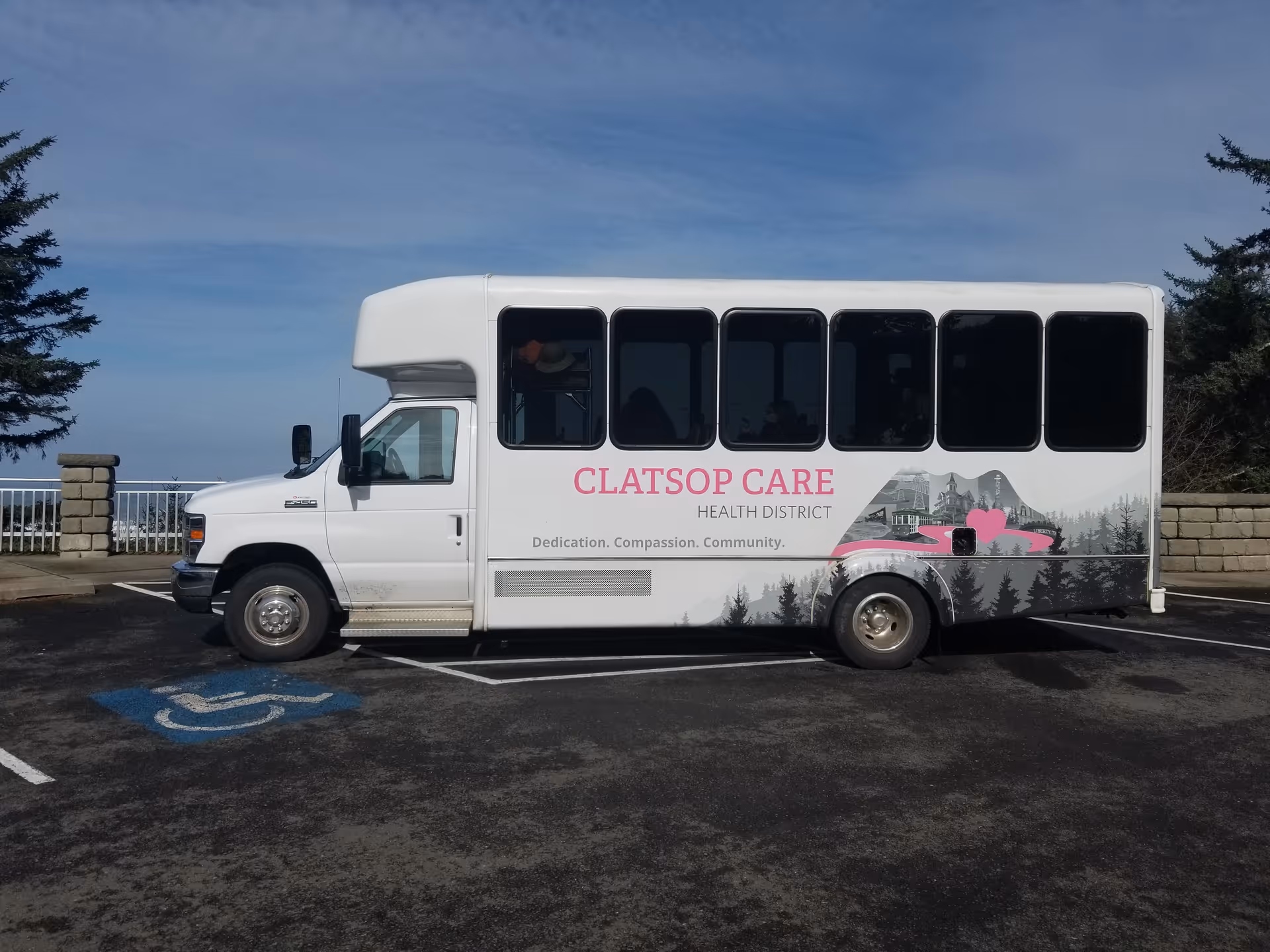 White Clatsop Care Health District shuttle bus parked in an outdoor lot with ocean and trees in the background.