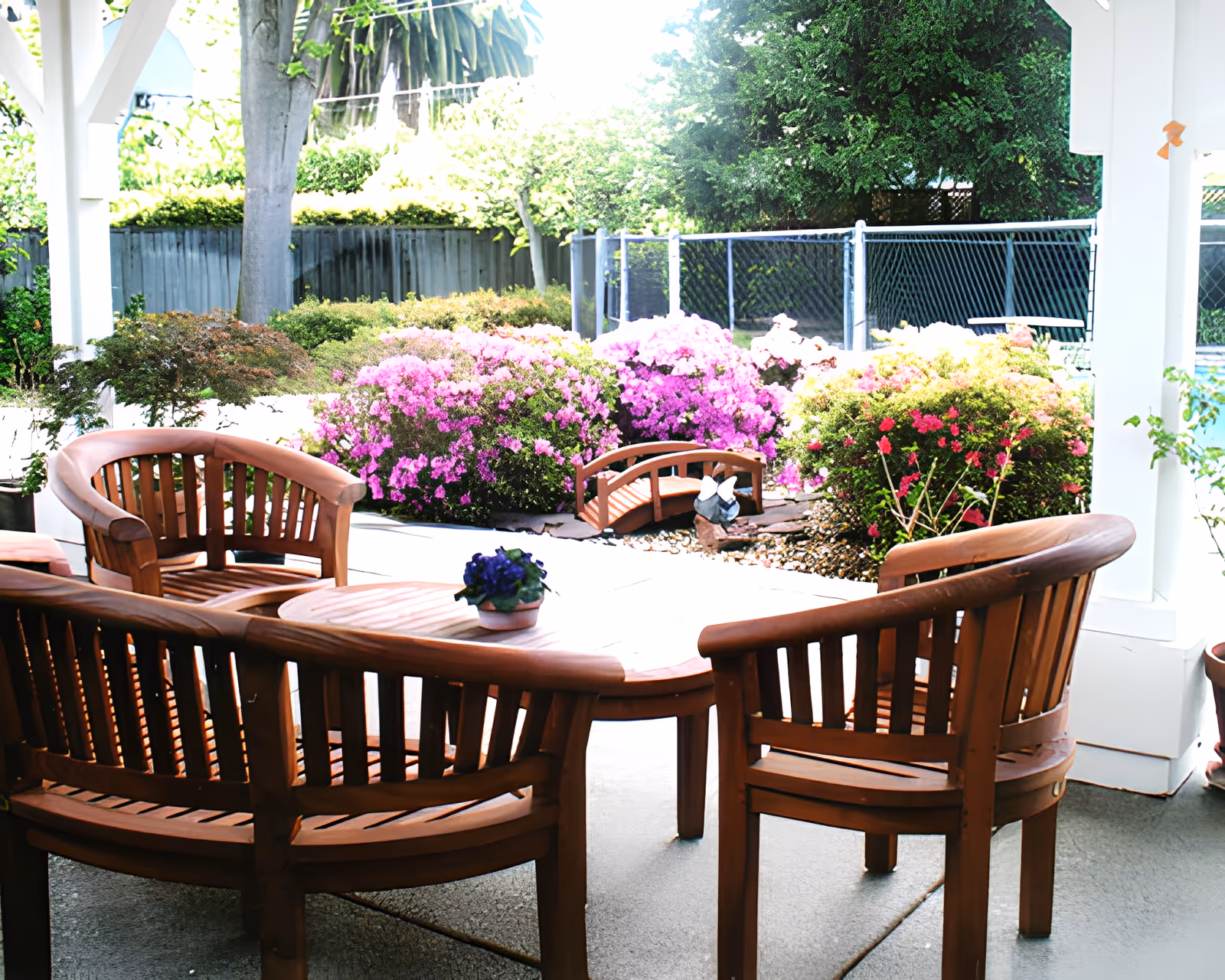Wooden patio table and chairs under a covered pergola overlooking colorful flowering shrubs and a small garden.