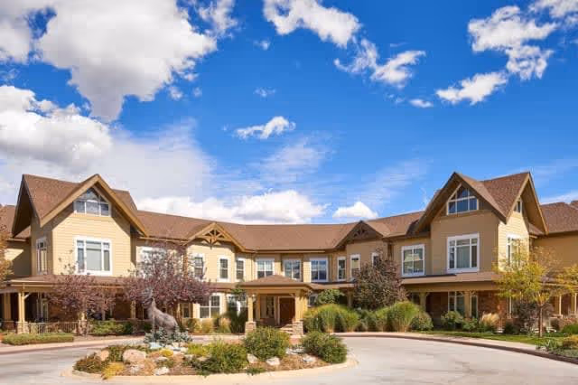 Exterior view of a large, two-story assisted living facility building with beige siding and brown roof. The building is surrounded by landscaped greenery, including bushes and small trees, under a bright blue sky with scattered clouds.