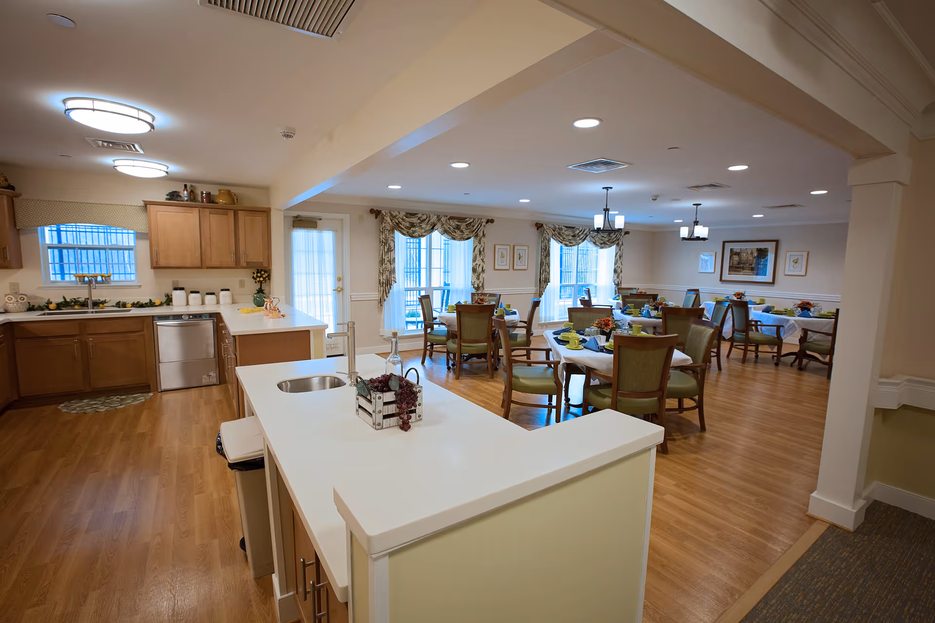 A spacious senior living facility dining area and kitchen with wooden floors, light wood cabinets, and white countertops. The kitchen features a sink, dishwasher, and various kitchen items. The dining area has several tables set with green chairs, tableware, and floral centerpieces. Large windows with decorative curtains allow natural light into the room.