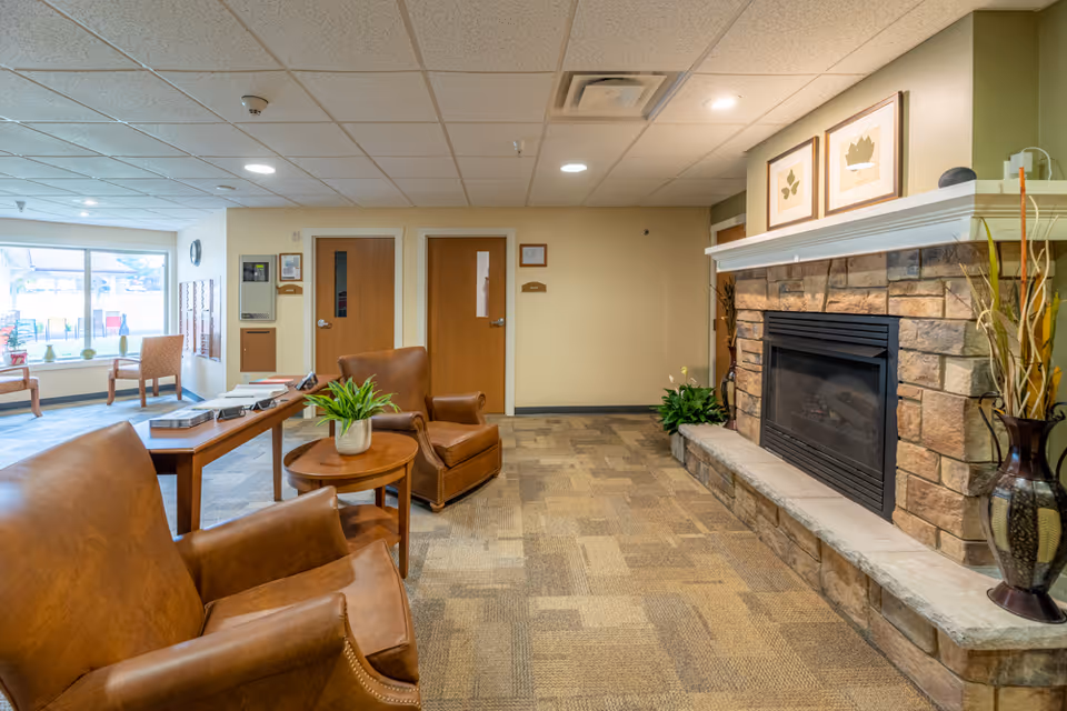 A cozy interior common area with two brown leather armchairs and a small round wooden table with a potted plant. There is a stone fireplace on the right wall with framed botanical prints above it. In the background, there are two wooden doors and a window with additional seating and natural light coming in.