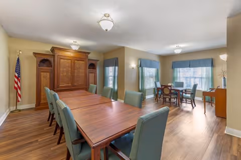 Communal dining room with long wooden tables and green upholstered chairs, large windows with blue valances, a wooden armoire, and an American flag.