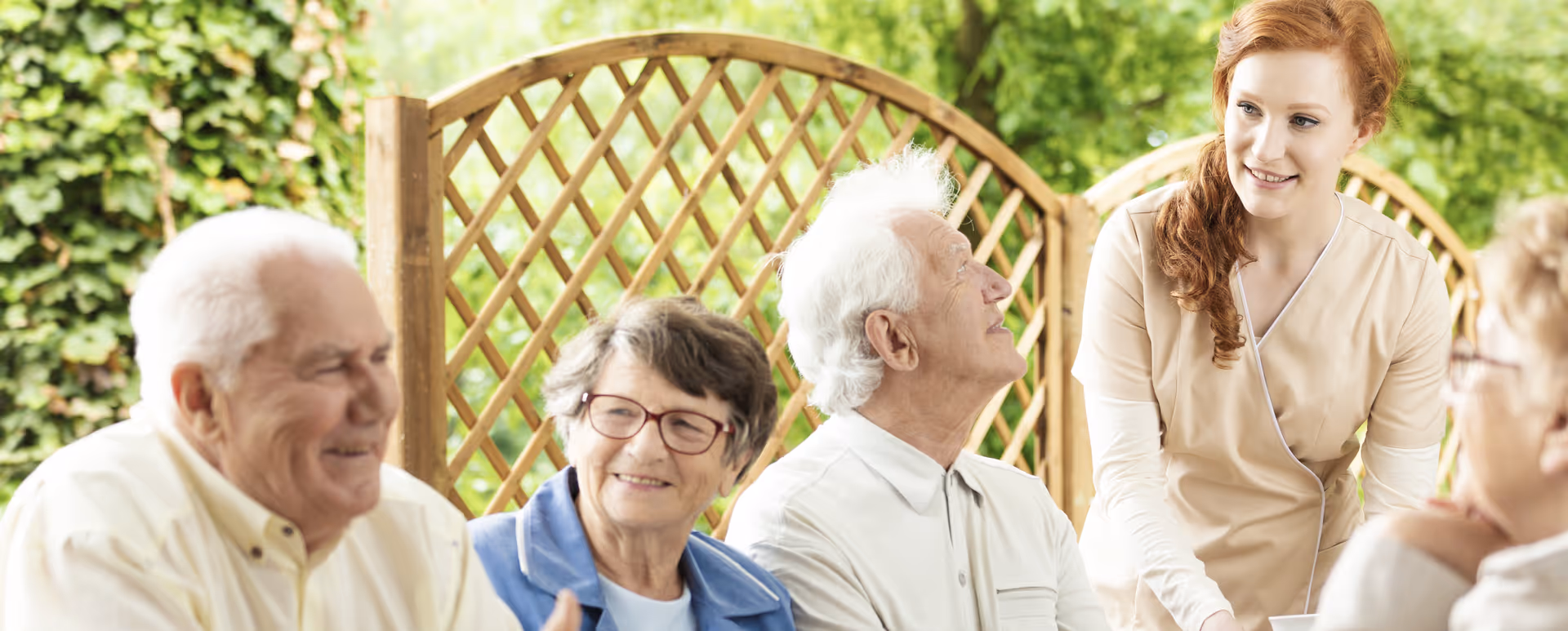 A group of elderly people sitting outdoors and smiling while a young female caregiver interacts with them. They are seated in front of a wooden lattice with greenery in the background.