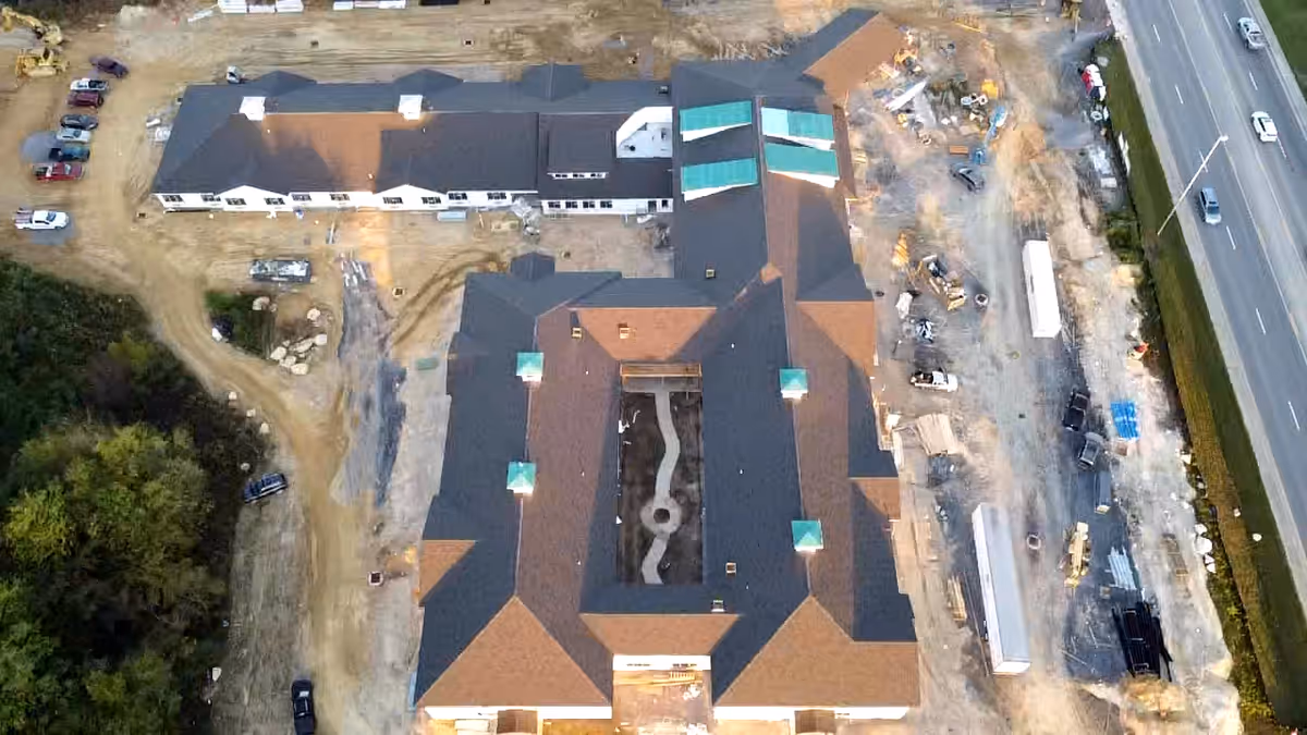 Aerial view of a large senior living facility under construction, showing the building's roof, surrounding construction equipment, dirt roads, and nearby highway with vehicles.
