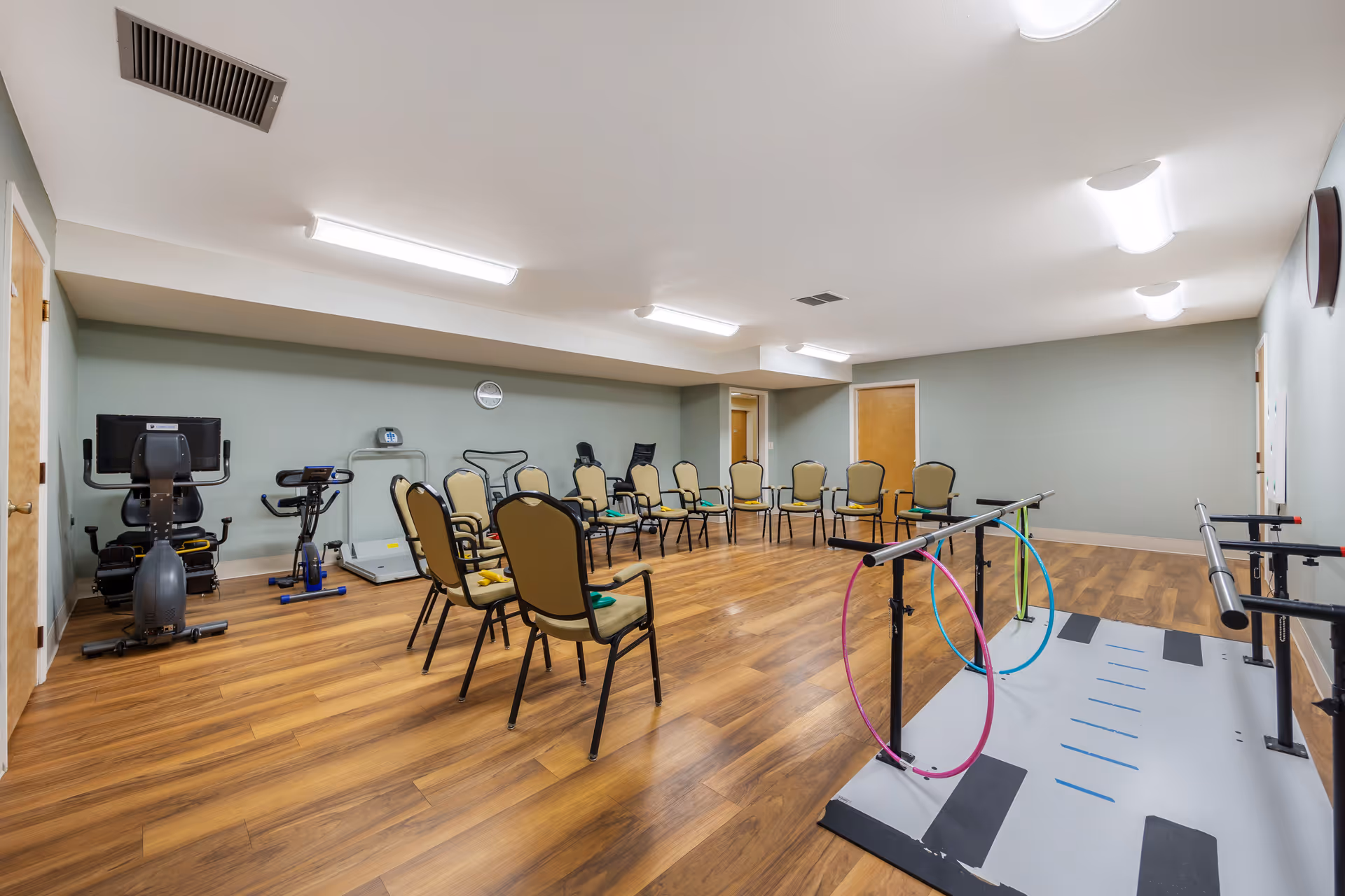 A spacious interior activity room with chairs arranged in a circle, exercise equipment, and parallel bars with colorful hoops on a wood floor.