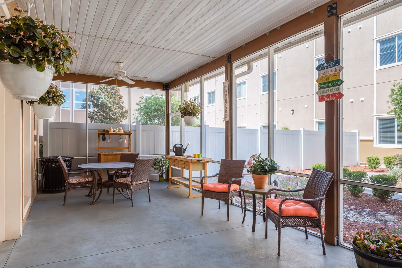 Covered screened patio with seating, tables, hanging plants, and potted flowers overlooking landscaped grounds.