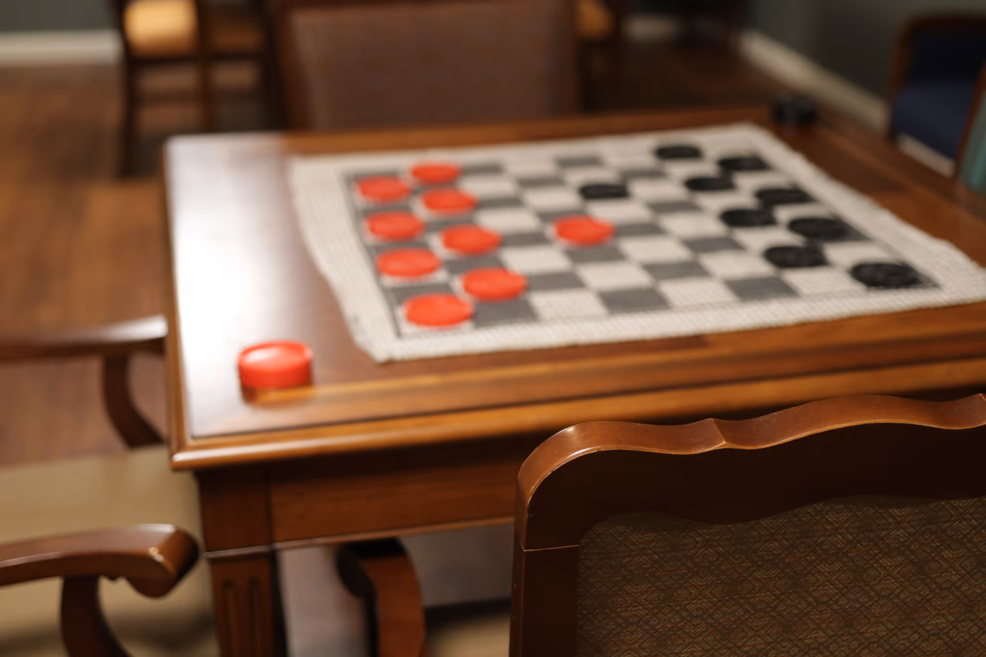 A wooden table with a checkers game set up on a checkered cloth. The checkers pieces are red and black. Surrounding the table are wooden chairs with cushioned seats and backs. The setting appears to be indoors with a wooden floor.