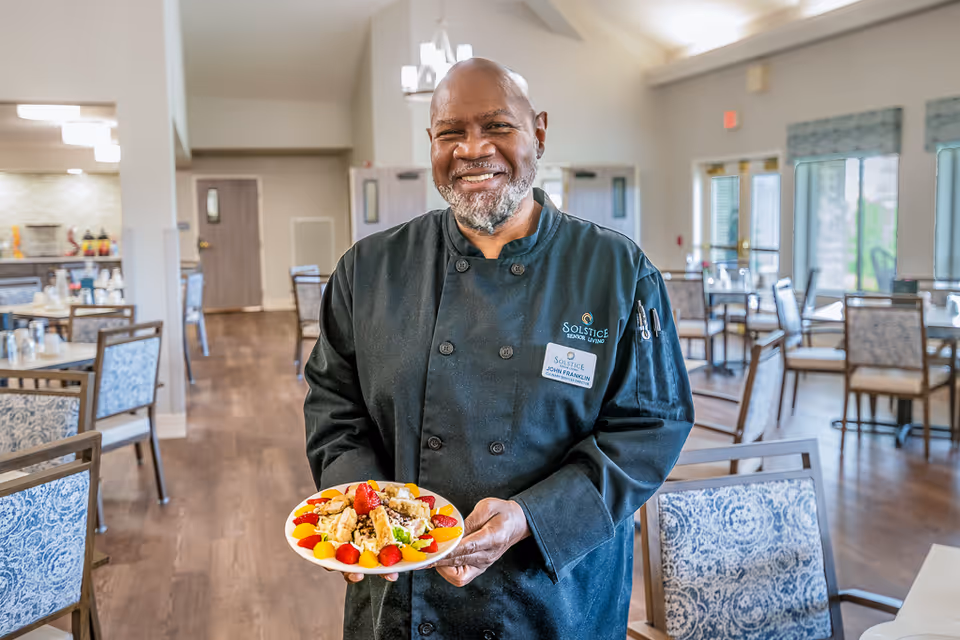A smiling chef wearing a black uniform and name tag that reads John Franklin stands in a dining room holding a plate of food garnished with strawberries and other fruits. The dining room has wooden floors, multiple tables and chairs with patterned upholstery, and large windows letting in natural light.