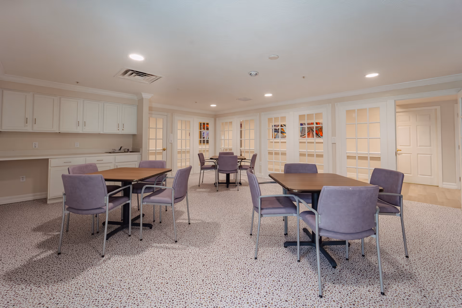 A clean, well-lit room with several square wooden tables surrounded by purple cushioned chairs. The room has white cabinetry along one wall and multiple glass-paneled doors. The carpet has a subtle dotted pattern, and the walls are painted a light beige color.