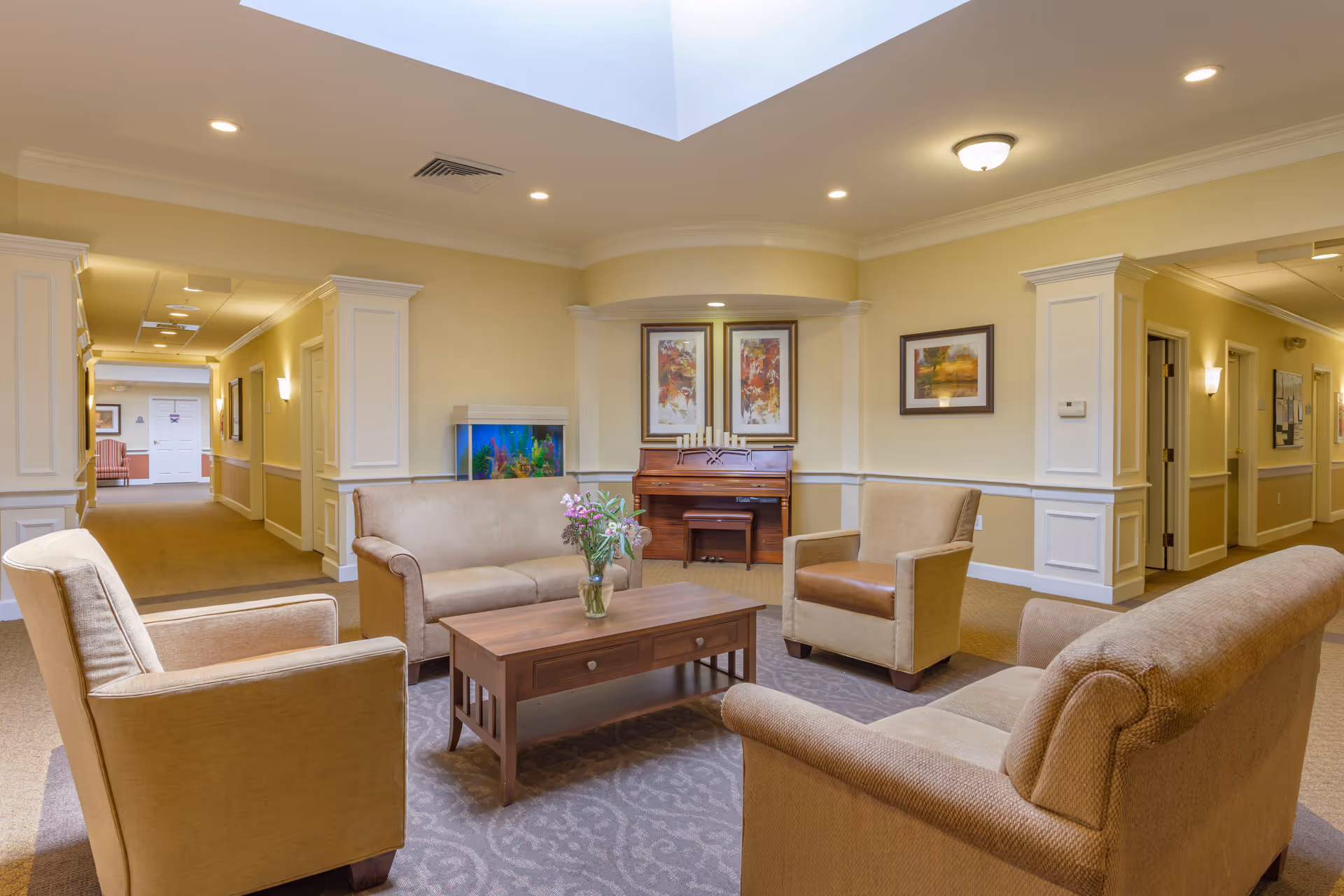 A cozy seating area in a senior living facility with beige armchairs and sofas arranged around a wooden coffee table with a vase of flowers. Behind the seating area is a piano and an aquarium, with framed artwork on the walls. The space is well-lit with ceiling lights and has a soft yellow wall color with white trim.