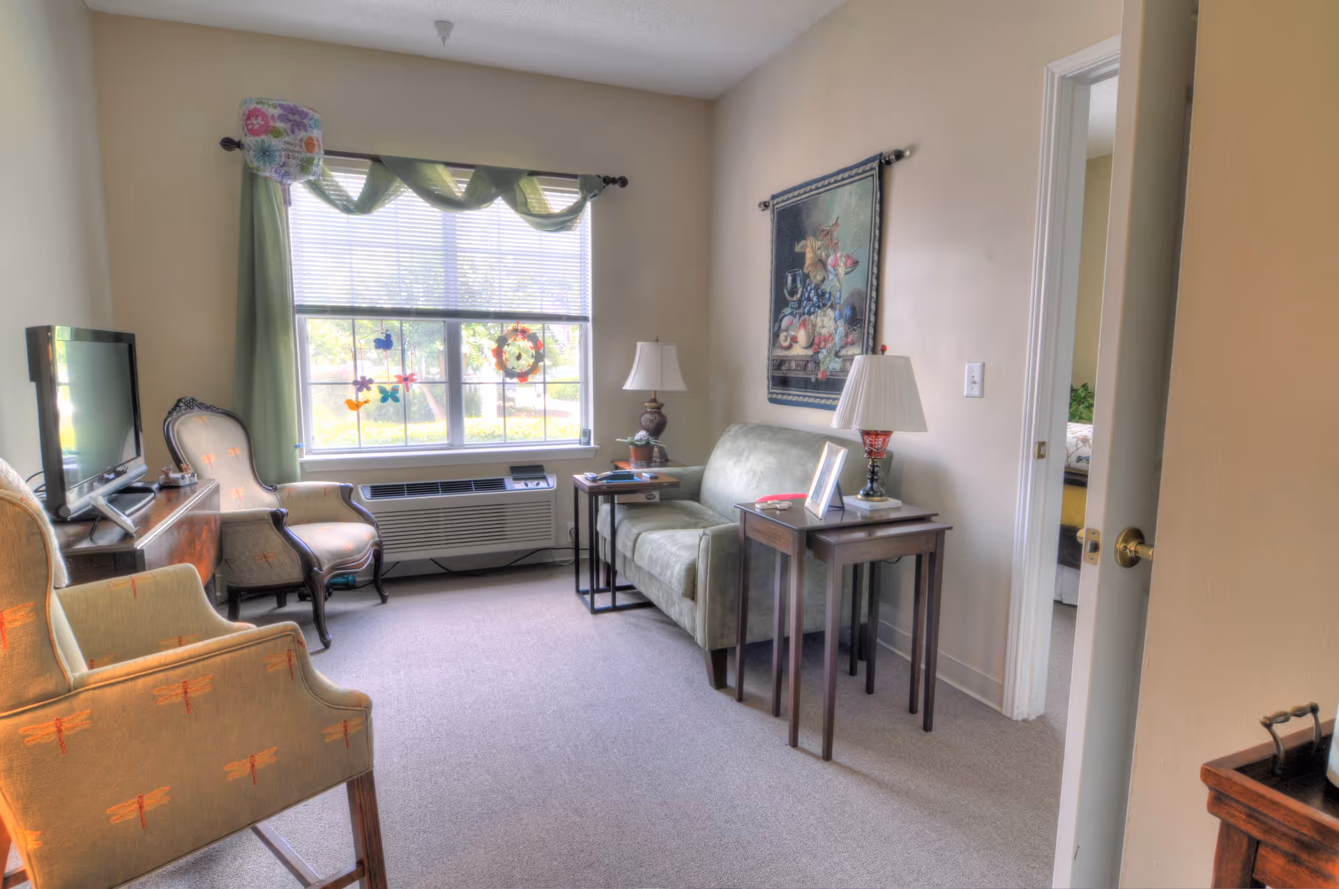 Small furnished living room with a sofa, armchairs, TV, side tables and a window with green curtains.