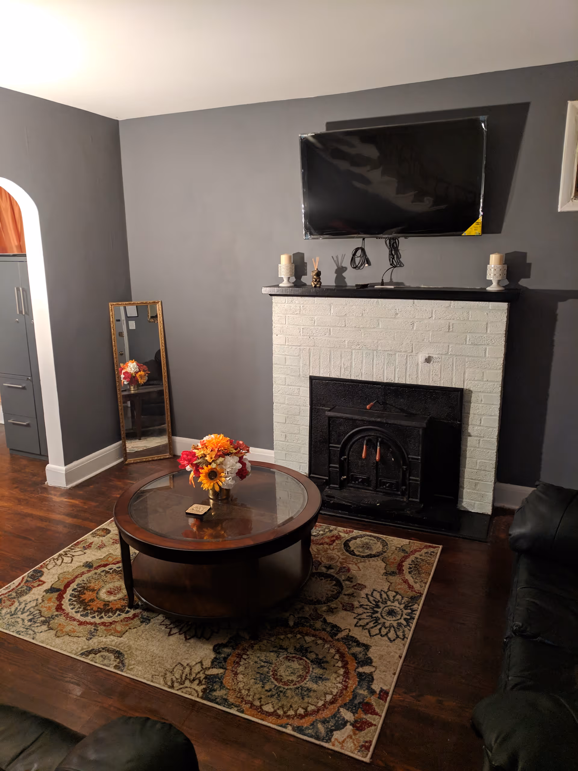 A cozy living room with dark gray walls, a white brick fireplace with a black wood stove insert, and a flat-screen TV mounted above the fireplace. In front of the fireplace is a round wooden coffee table with a glass top, decorated with a vase of colorful flowers. A patterned area rug lies beneath the coffee table. To the left, there is a tall mirror leaning against the wall, reflecting part of the room. Dark leather furniture is partially visible on the right and bottom edges of the image.