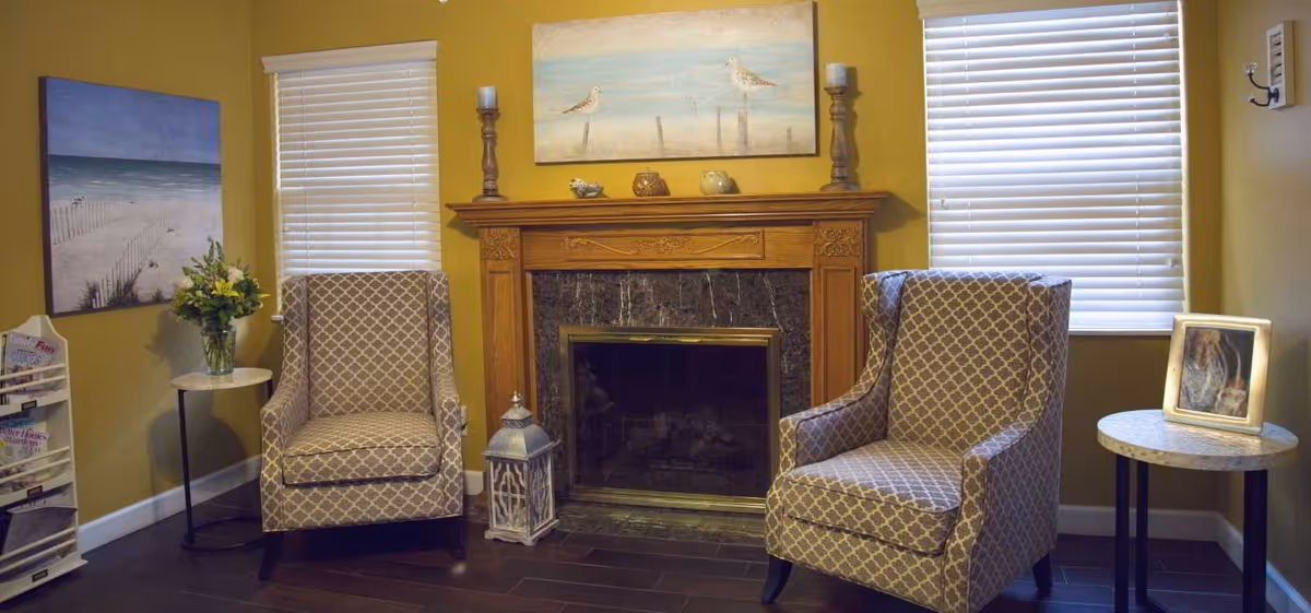 A cozy sitting area with two patterned armchairs placed on either side of a wooden fireplace with a dark marble surround. Above the fireplace is a painting of seagulls on a beach. Two windows with white blinds flank the fireplace. A small round table with a vase of flowers is next to the left chair, and another round table with a framed photo is next to the right chair. A magazine rack is visible on the far left side of the room.