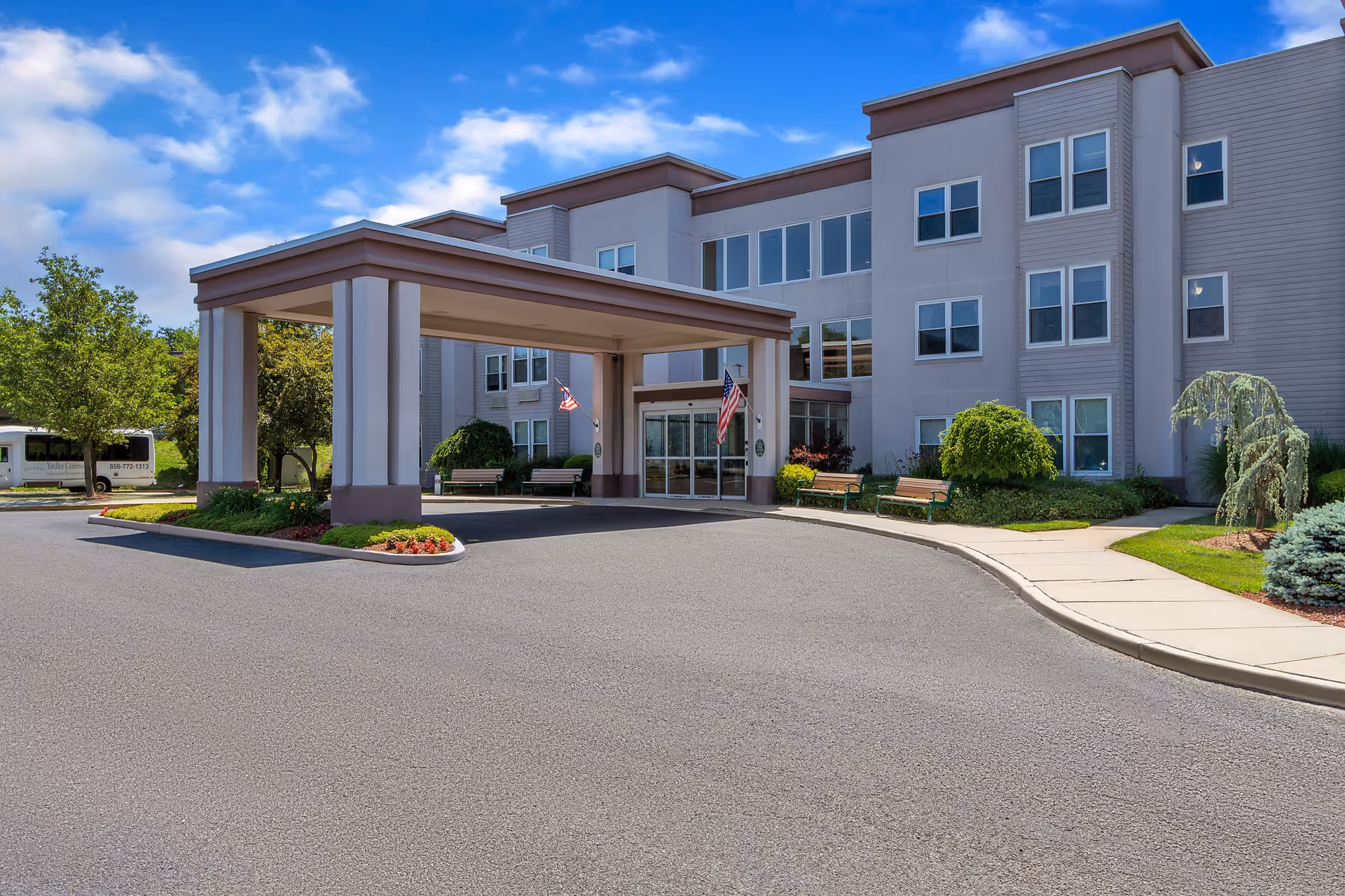 Exterior view of Seaton Voorhees senior living facility showing the main entrance with a covered drop-off area, benches, landscaping with bushes and trees, and a clear blue sky with some clouds.