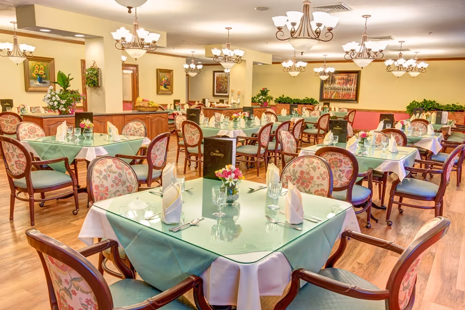 A bright and elegant dining room with multiple tables covered in light green tablecloths and white napkins. Each table is set with glassware, silverware, and small floral centerpieces. The chairs have wooden frames with floral upholstery. The room is decorated with framed paintings, plants, and warm lighting from chandeliers hanging from the ceiling.