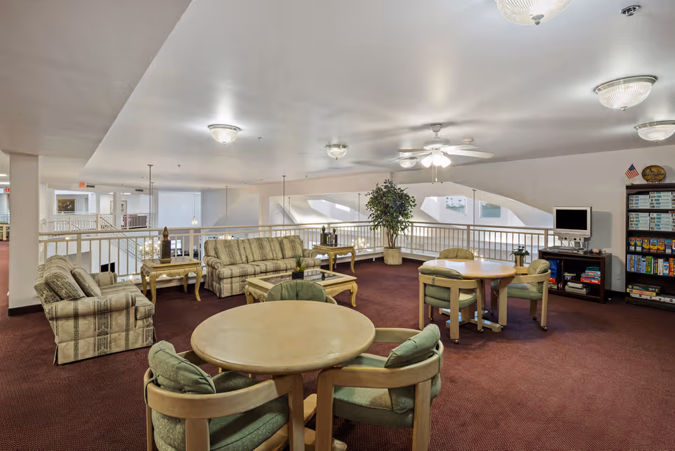 A senior living community common area with round tables and cushioned chairs, two striped sofas, a coffee table, side tables with decorative items, a potted plant, a TV on a stand, and a bookshelf filled with board games. The space has carpeted floors, white walls, ceiling lights, and a ceiling fan.