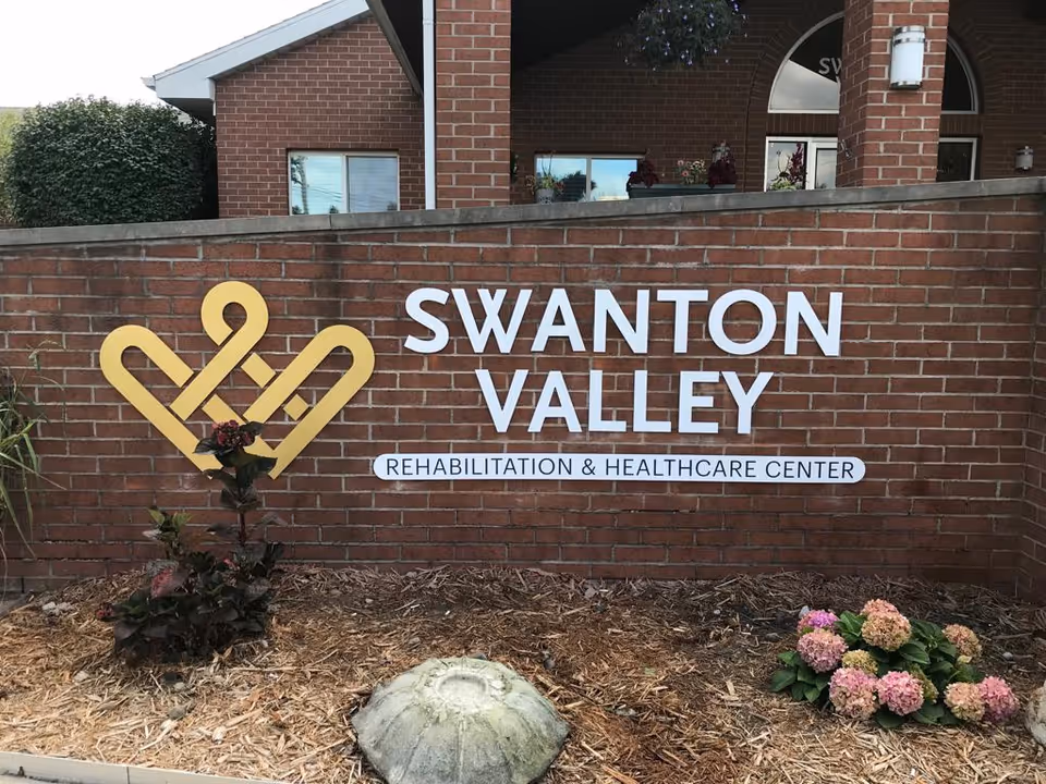 Brick wall with a yellow logo and white text reading 'Swanton Valley Rehabilitation & Healthcare Center' with some plants and flowers in front of the wall and part of a building visible behind the wall.