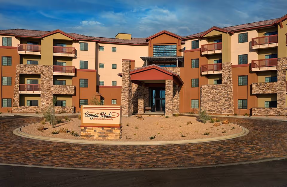 Front exterior view of Canyon Winds Assisted Living & Memory Care building with a circular driveway, desert landscaping, and a sign that reads 'Canyon Winds Retirement Community'. The building has multiple floors with balconies and a mix of stone and stucco facade under a partly cloudy sky.