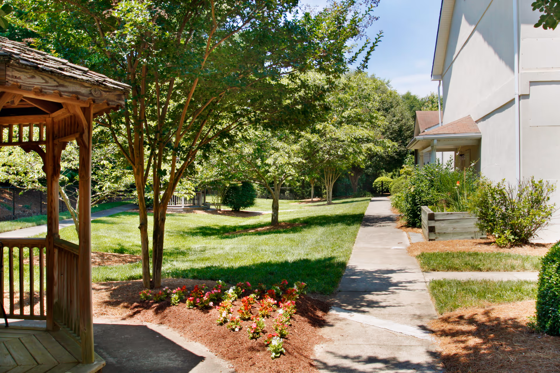 A sunny outdoor garden area with a wooden gazebo on the left, a paved walkway, green grass, trees, and flower beds near a light-colored building.