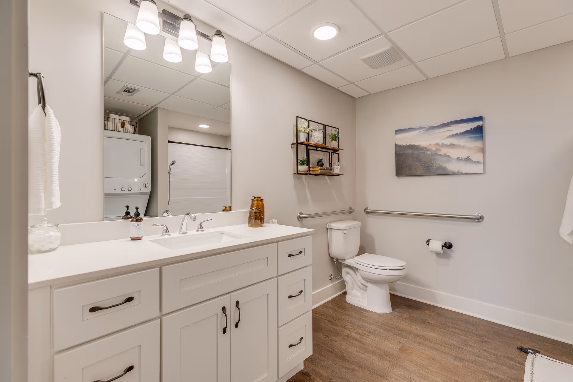 A clean and modern bathroom featuring a white vanity with a large mirror and three light fixtures above it. The bathroom has a toilet with grab bars on the walls for accessibility, a wall-mounted shelf with decorative items, and a framed landscape picture on the wall. The floor is wood-style, and a stacked washer and dryer unit is visible in an adjacent laundry area.