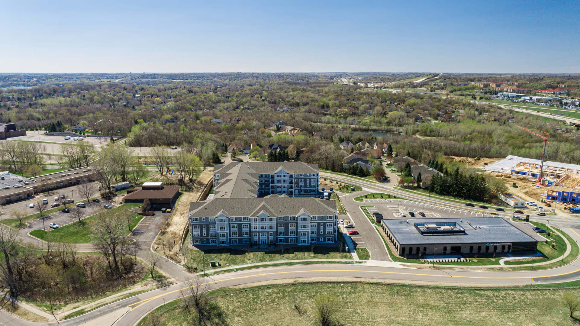 Aerial view of a senior living facility named Havenwood of Burnsville, showing multiple buildings surrounded by parking lots, roads, and green wooded areas under a clear sky.