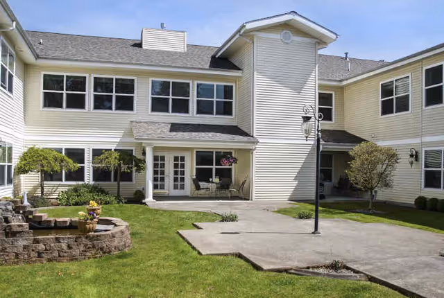 Exterior view of a two-story assisted living facility with beige siding, multiple windows, a small patio with chairs and a table, a lamp post, and a well-maintained lawn with some small trees and plants.
