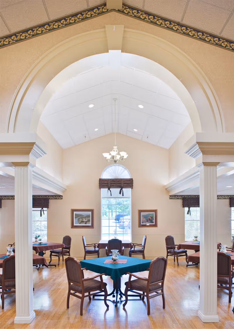 Interior view of a dining room in Canterbury Center featuring multiple tables covered with tablecloths and surrounded by wooden chairs. The room has high vaulted ceilings with a chandelier, large windows with brown valances, framed artwork on the walls, and polished wooden floors.