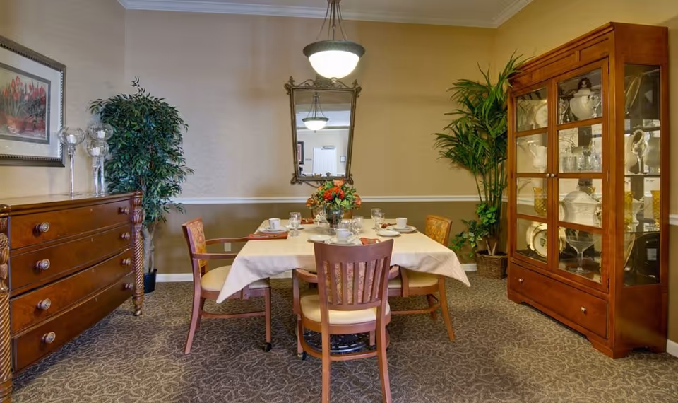 A dining room with a rectangular table covered with a white tablecloth, set with cups, glasses, and plates. Four wooden chairs with cushioned seats surround the table. The room features a wooden dresser on the left, a glass-fronted wooden cabinet displaying dishes on the right, two potted plants, a framed floral picture on the left wall, and a decorative mirror hanging on the back wall under a ceiling light fixture.