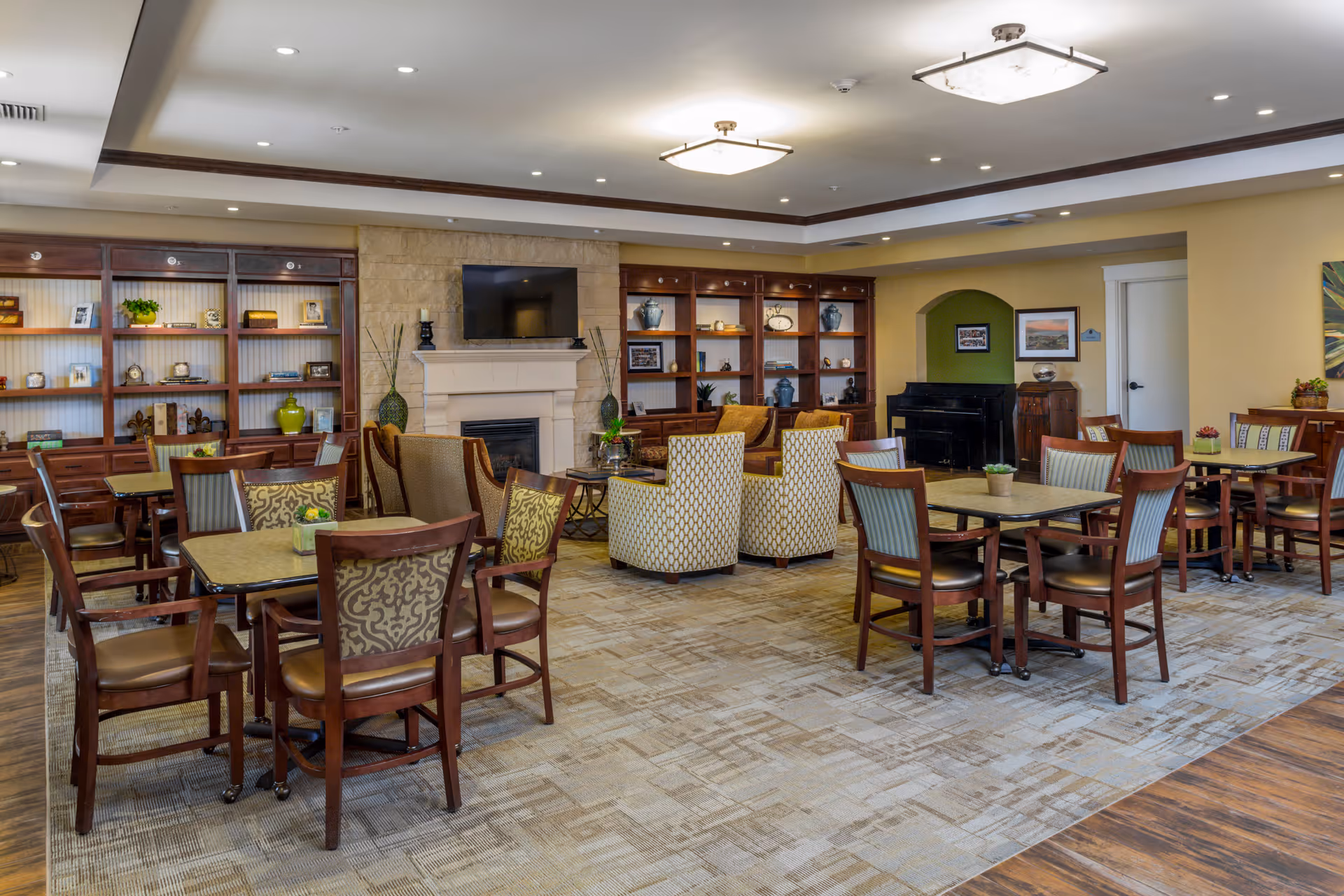 A spacious and well-lit common area with multiple tables and chairs arranged for seating. The room features a fireplace with a mounted TV above it, built-in wooden shelves with decorative items, and comfortable armchairs arranged around a coffee table. The walls are painted in warm tones, and there is a piano in the corner near a doorway.
