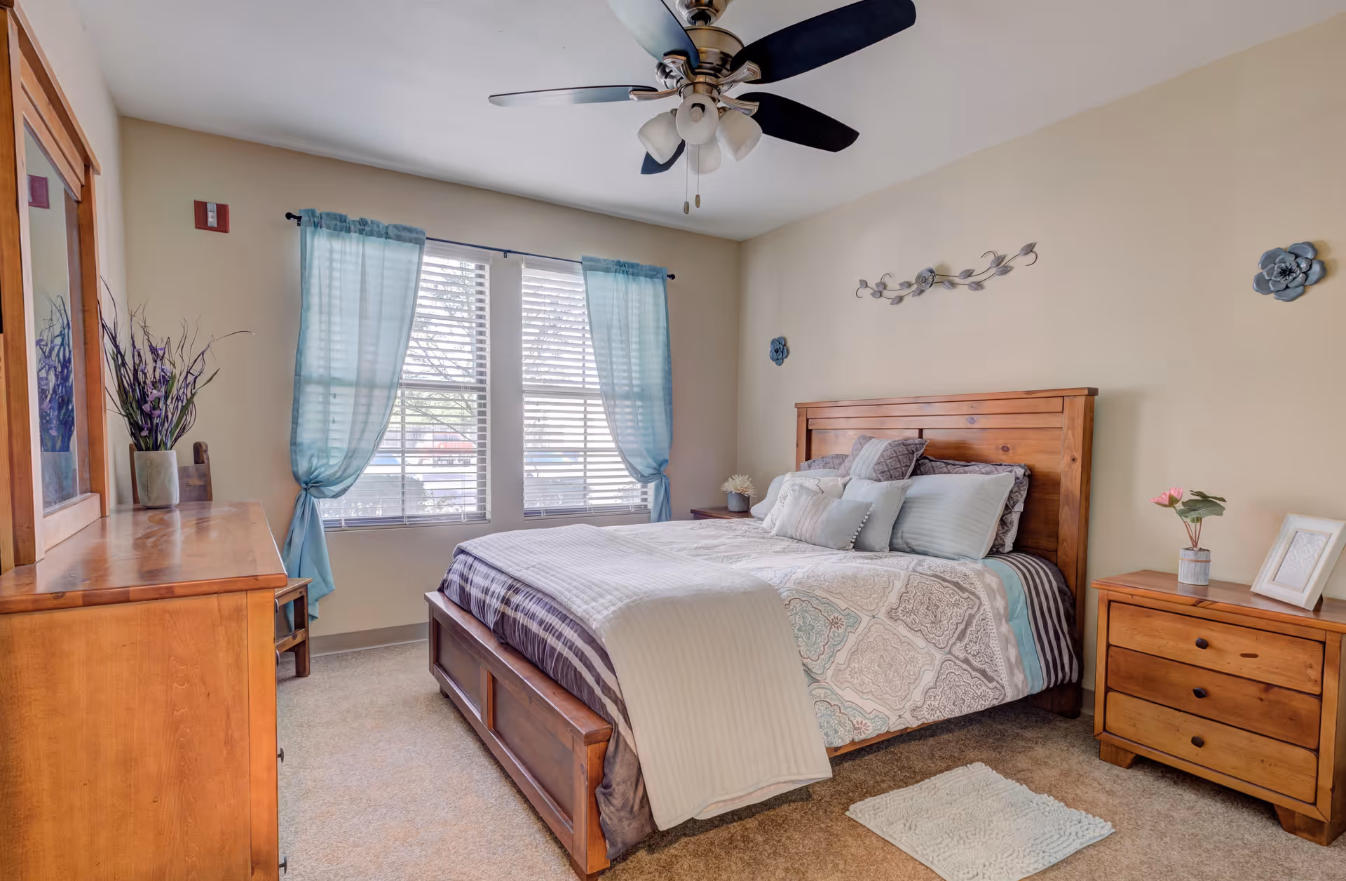 A cozy bedroom with a wooden bed frame and matching wooden dresser and nightstand. The bed is neatly made with patterned bedding and multiple pillows. Light blue curtains frame two large windows letting in natural light. A ceiling fan with lights is mounted on the ceiling. Decorative wall art and a small plant on the nightstand add a homey touch.